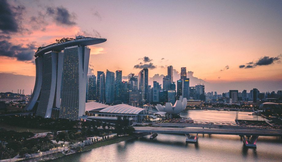 Aerial view of Singapores Marina Bay Sands and skyline at sunset. The iconic hotel towers over the cityscape with the ArtScience Museum nearby. The sky is a mix of orange and purple hues, reflected on the calm waters of the bay.