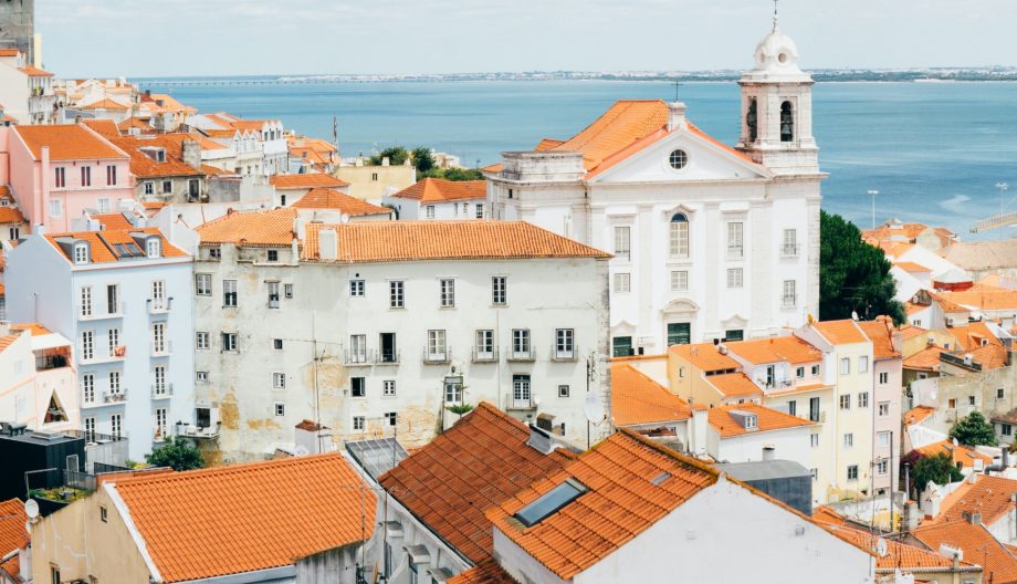 A scenic view of a coastal city with red-tiled rooftops in the foreground. A white church with a bell tower stands prominently among the buildings. The ocean and a clear blue sky with scattered clouds are visible in the background.