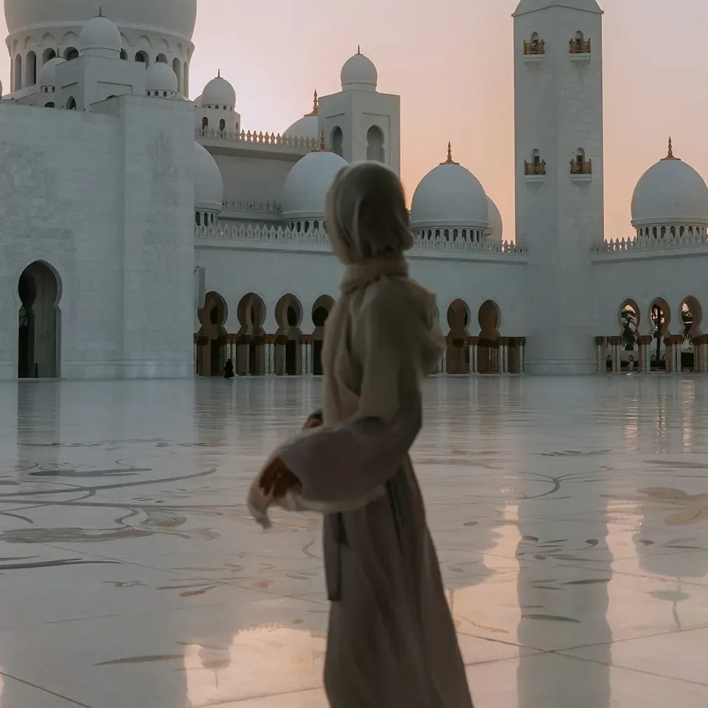 A person in modest clothing stands on a reflective marble courtyard at the Sheikh Zayed Grand Mosque, with white domes and minarets in the background during sunset.