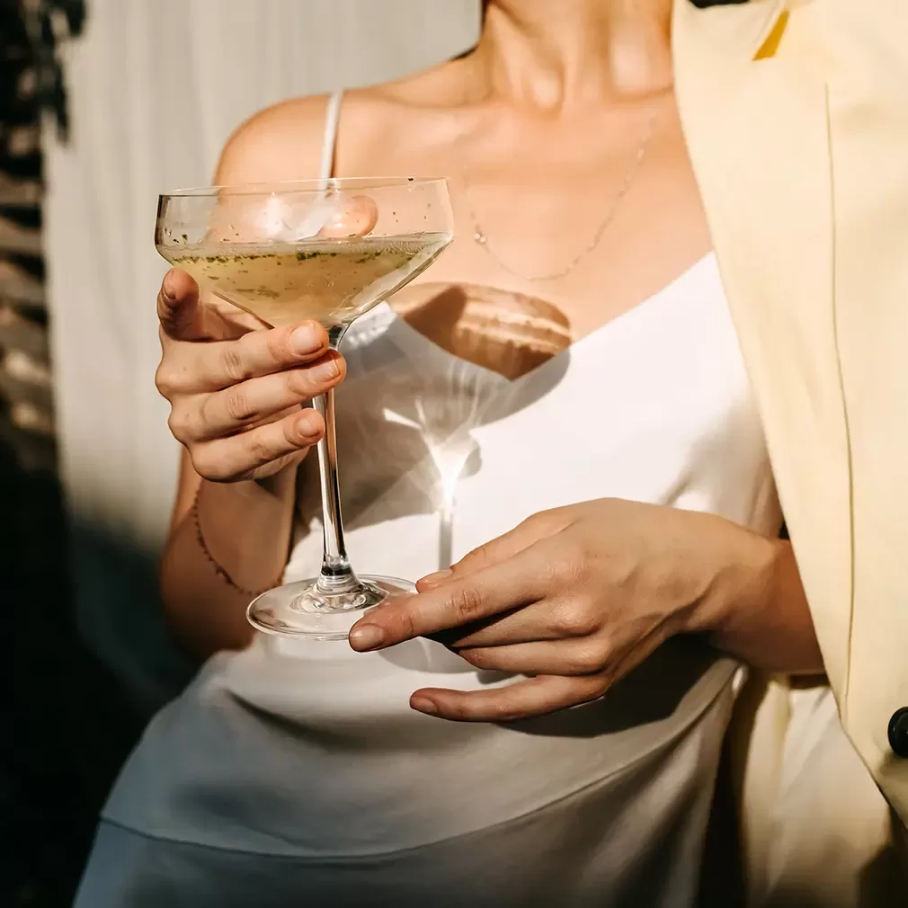 A woman in a white satin dress holds a cocktail glass filled with a light-colored drink, standing next to someone in a light-colored jacket. Only her torso and hands are visible.