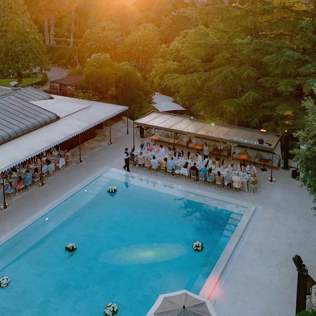Aerial view of an outdoor dinner event beside a pool at sunset, with guests seated at long tables under awnings, surrounded by trees and greenery.