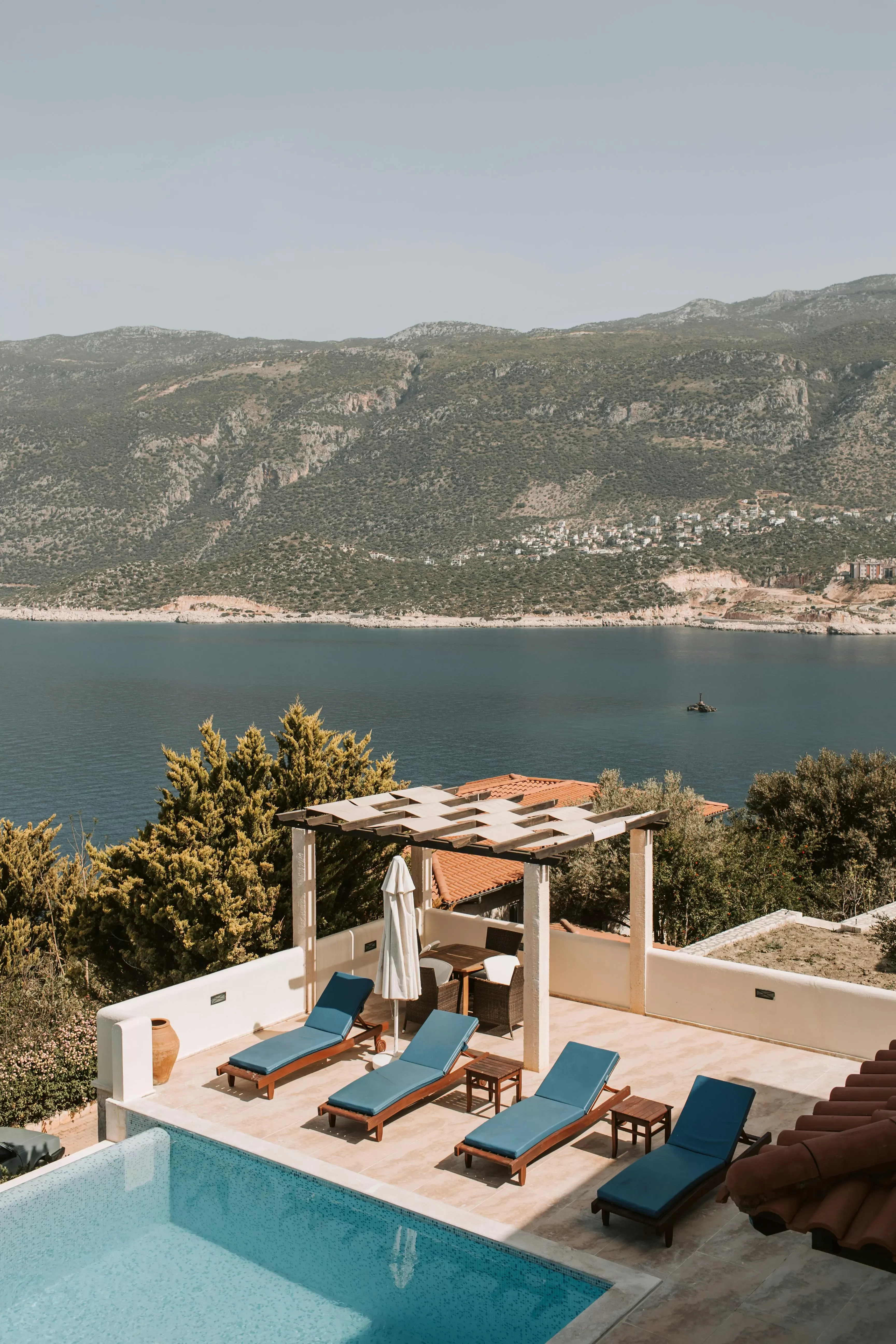 A terrace with blue lounge chairs by a pool overlooks a calm sea and green mountains in the background under a clear sky.