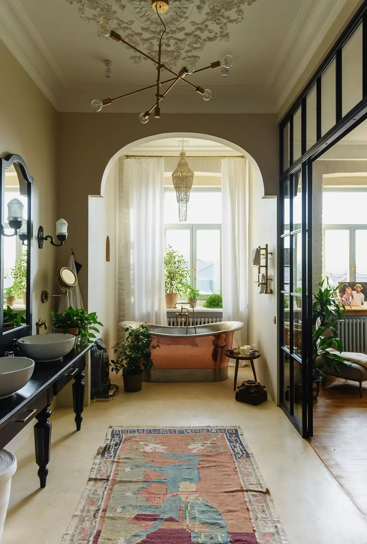 Elegant bathroom with a freestanding tub, curtains, plants, a patterned rug, double sinks, and large windows letting in natural light. A chandelier and ornate ceiling details add a luxurious touch.