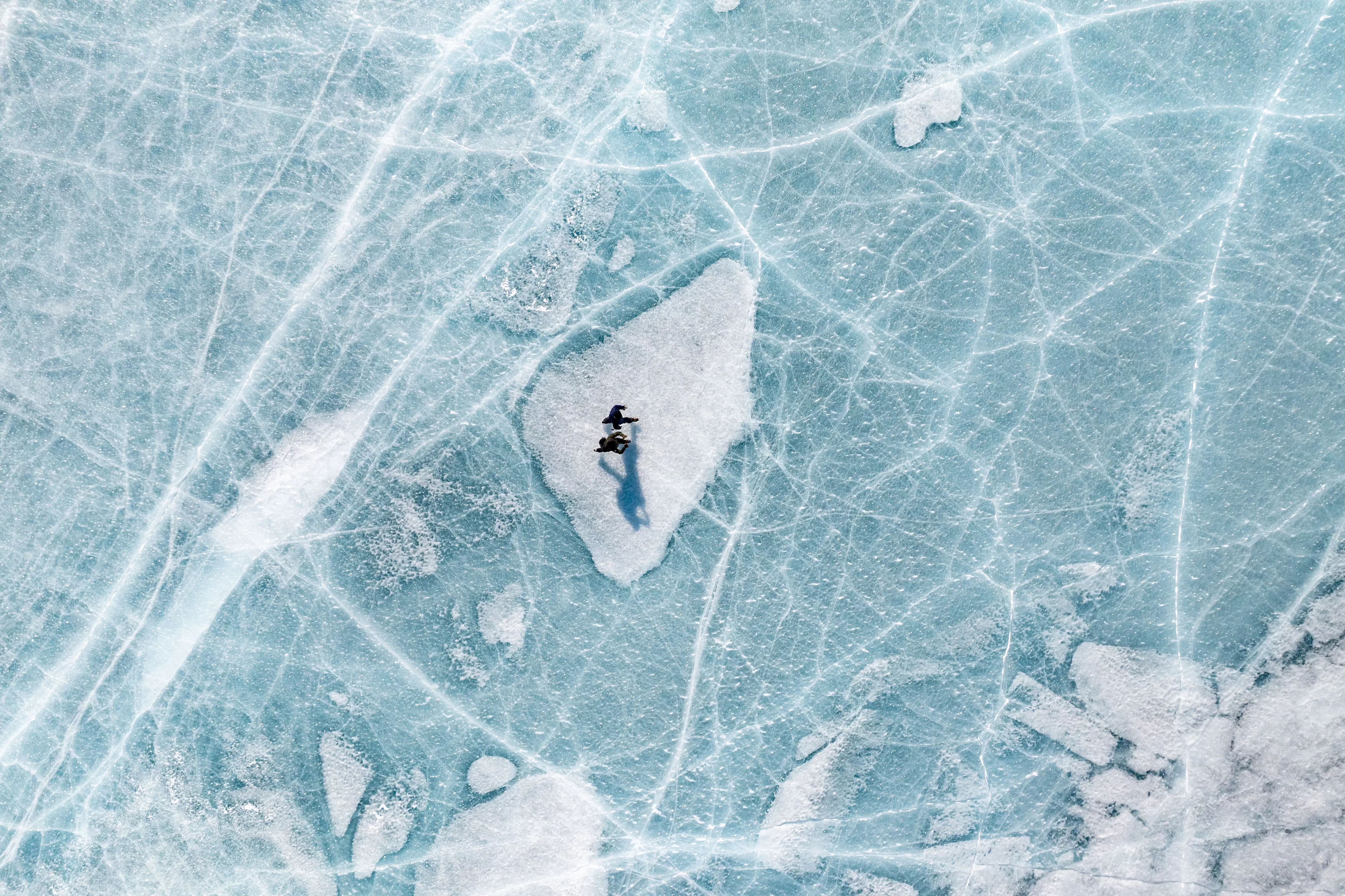 A person stands on a white patch of ice, surrounded by blue cracked and textured frozen lake surface, casting a long shadow. The scene is viewed from above.
