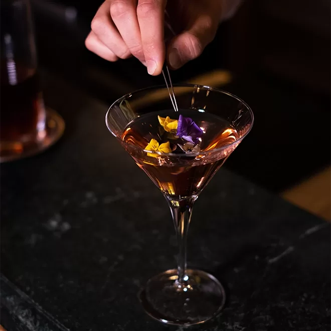 A hand delicately places edible flowers into a martini glass filled with a pink cocktail, set on a dark countertop.