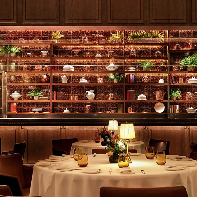 A warmly lit restaurant with round tables set for dining, featuring a decorative backlit shelving unit displaying plants, teapots, vases, and books, creating an elegant and cozy atmosphere.