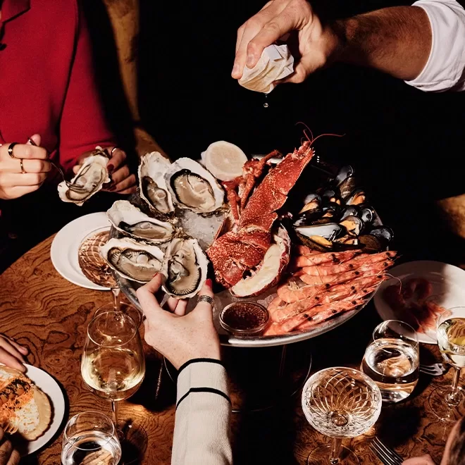 A platter of assorted seafood including oysters, lobster, mussels, and shrimp sits on a table with several glasses of white wine, as people reach in to enjoy and one hand squeezes lemon over the seafood.