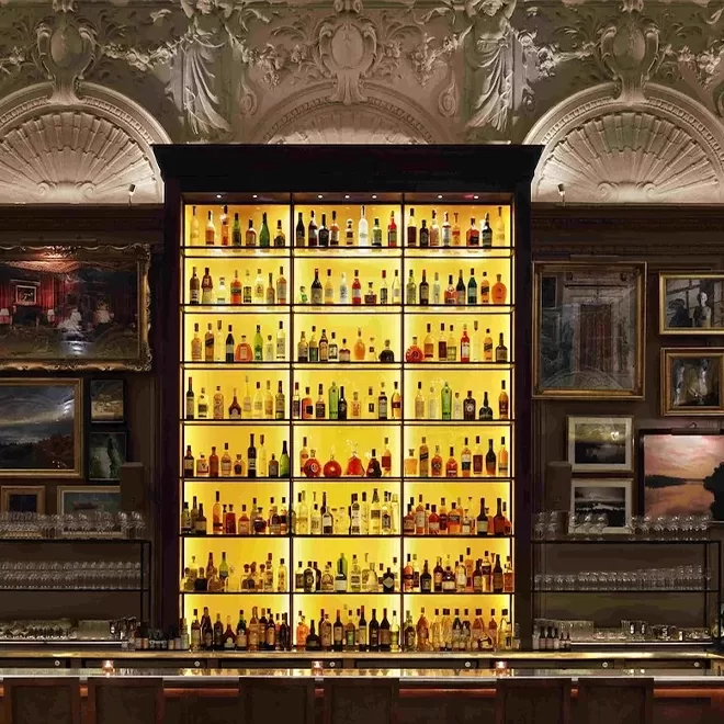 A backlit bar shelf filled with various bottles of liquor is framed by ornate carvings and surrounded by framed artwork. The overall ambiance is luxurious and classic.