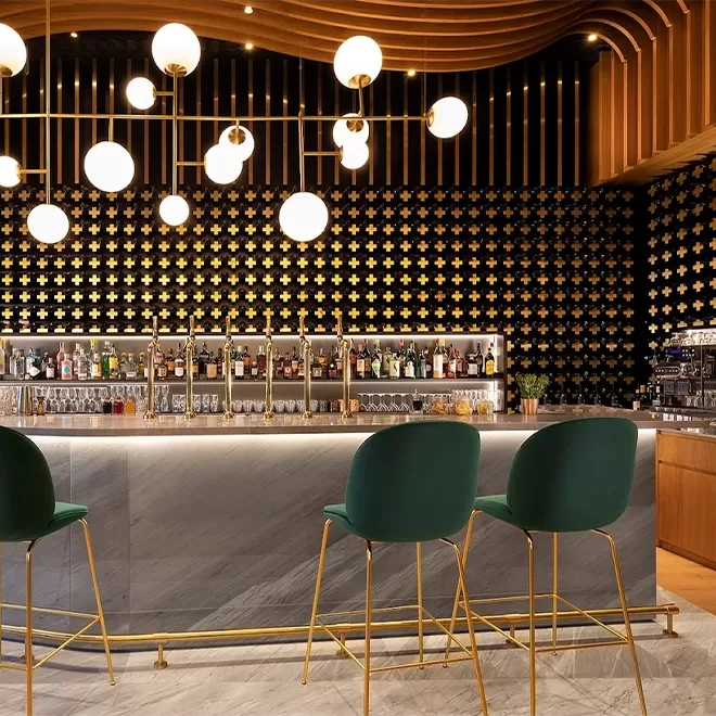 Modern bar with a marble counter, three green velvet barstools with gold legs, illuminated shelves of bottles, and decorative gold and black patterned wall, with round globe pendant lights overhead.