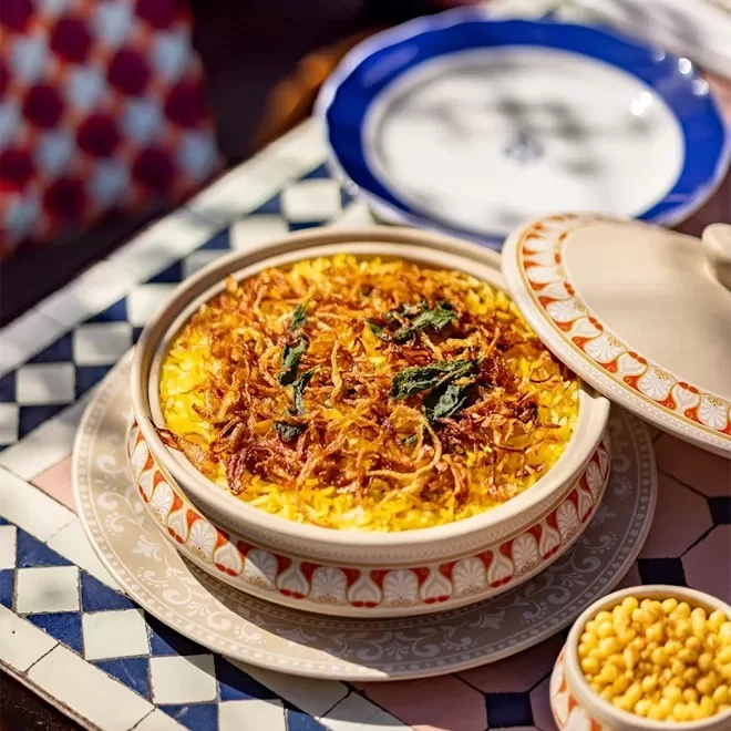 A decorative ceramic bowl filled with saffron rice topped with crispy fried onions and herbs sits on a patterned tray, with a matching lid and an empty blue-and-white plate in the background.
