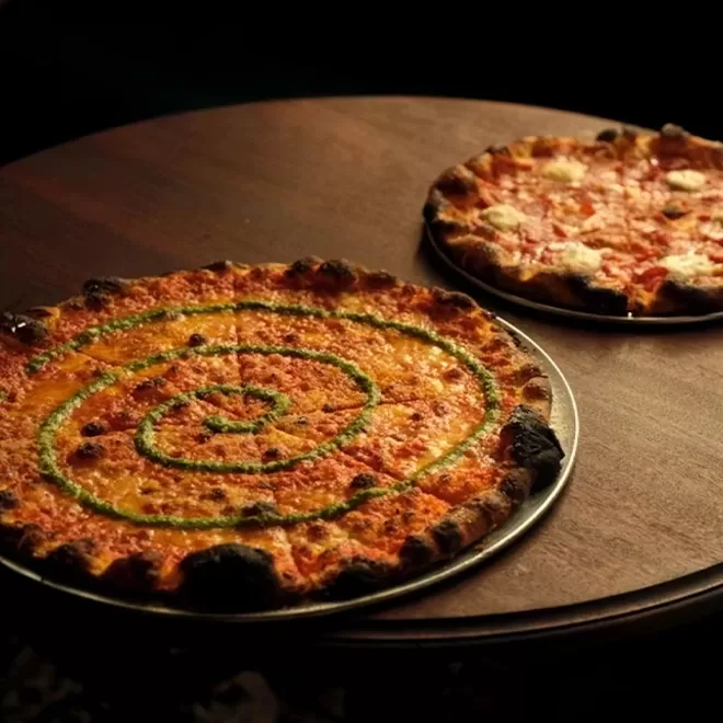 Two pizzas on a round wooden table; one pizza has a green spiral sauce pattern, while the other appears to be a classic cheese pizza with some dark, crispy edges.