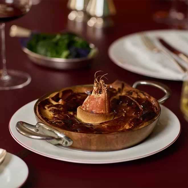 A round copper pan on a white plate holds a baked seafood dish, topped with a whole shrimp standing upright. The table is set with cutlery, a wine glass, and a small bowl of greens in the background.