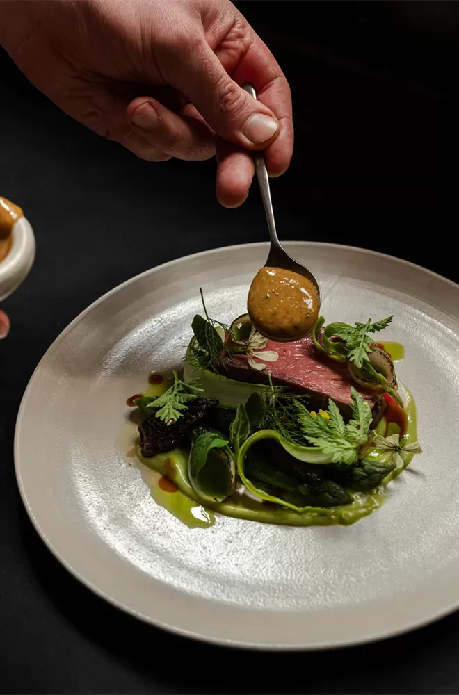 A person garnishes a gourmet dish with a spoonful of sauce. The plate features a piece of cooked meat, green vegetables, and herbs, presented artistically against a dark background at Evelyn’s Table, Soho, London.