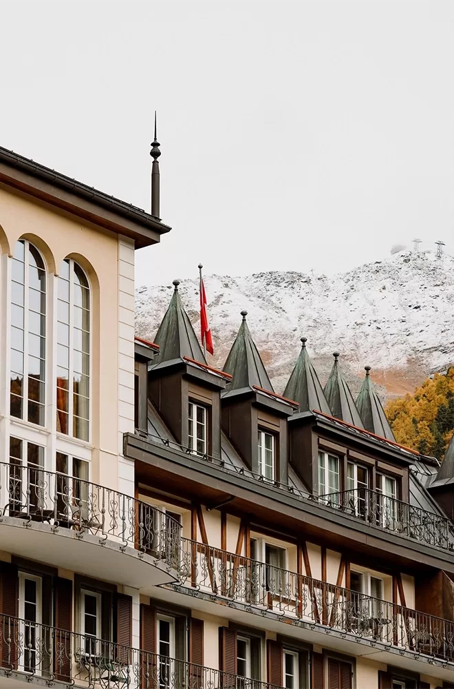A charming building with ornate balconies and triangular rooftops sits against a snowy mountain backdrop. A red flag is visible atop the building, contrasting with the clear sky and autumnal trees at Mont Cervin Palace, Zermatt.