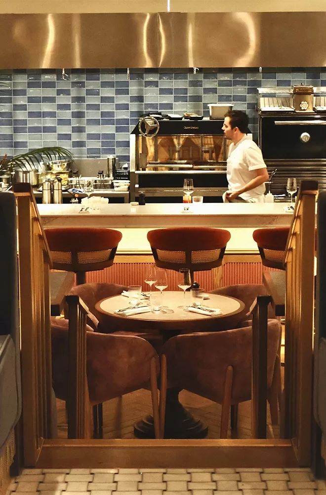 A restaurant interior with a set table and brown chairs in the foreground. Behind, a chef in a white uniform works in an open kitchen with blue tiled walls and various kitchen appliances at Three Darlings, London.