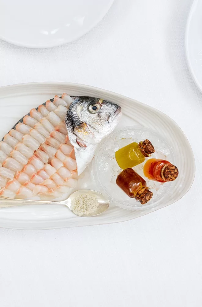 A plated gourmet dish featuring a fish head with carefully arranged sliced fish flesh. Accompanied by colorful small bottles on ice and a spoon with a powder-like substance, all presented on a white oval plate against a white background at Gaia, London.