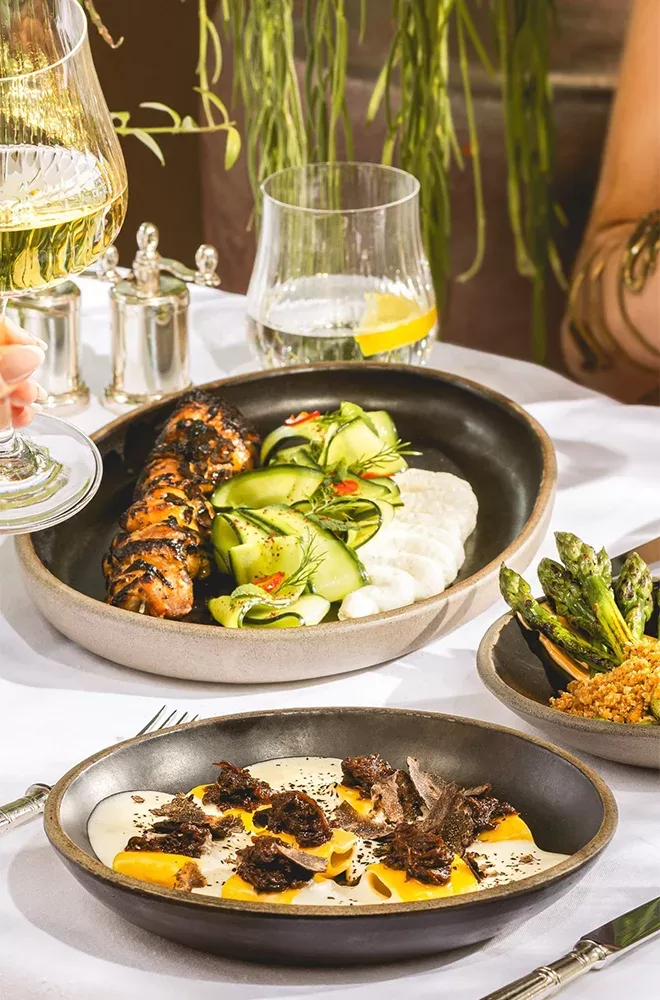 Elegant table setting with dishes featuring grilled skewers, cucumber salad, and creamy sauce pasta topped with truffles. Sides include quinoa with asparagus. Glasses of white wine and water are also on the table, set against a backdrop of greenery at Bacchanalia, London.
