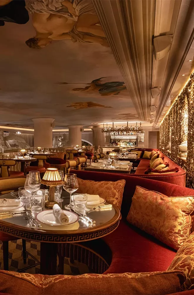 Luxurious restaurant interior with opulent decor, including plush red seating, elegantly set tables with white linens, and flanked by a sparkling gold wall. The ceiling features classical paintings, and a well-stocked bar is visible in the distance at Bacchanalia, London.