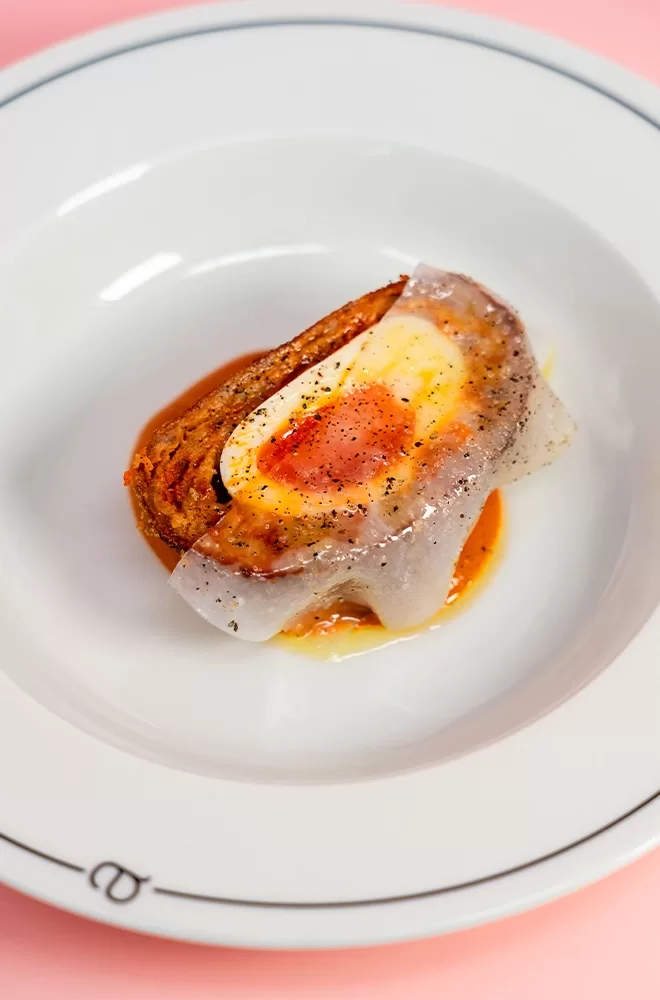 A gourmet dish featuring a soft-boiled egg with a runny yolk on top of a browned pastry slice, drizzled with sauce and sprinkled with black pepper, served on a white plate with a pink background at Three Darlings, London.