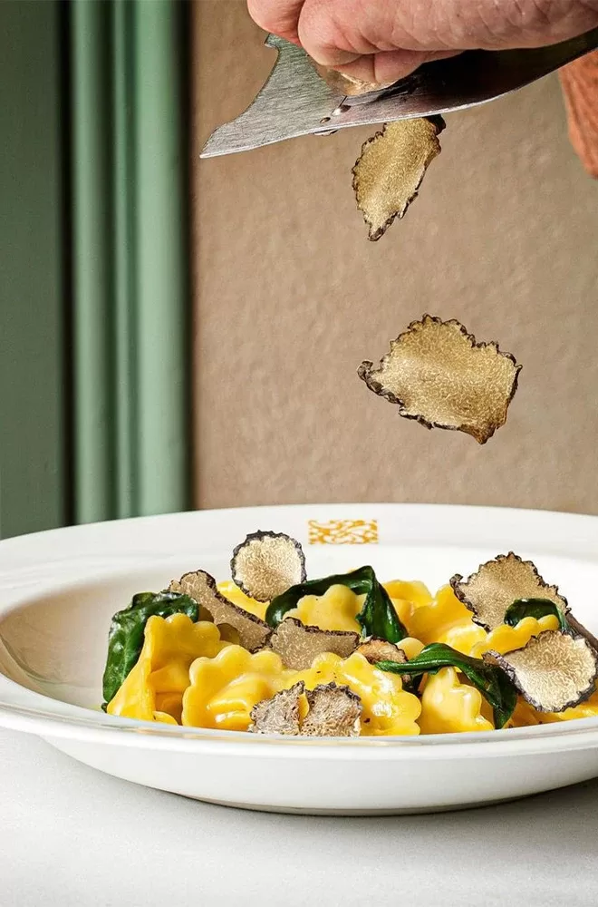 A bowl of pasta is topped with thin slices of truffle being shaved from a hand-held slicer. The dish is garnished with spinach leaves, and the scene is set against a soft-focus background at Daphne's, London.