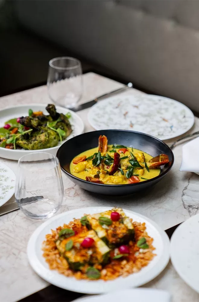 A table with assorted dishes, including a bowl of yellow curry with vegetables, a plate of roasted vegetables, and a dish with baked entrees garnished with greens and radishes. Two empty glasses and cutlery are also on the table at Kutir, London.