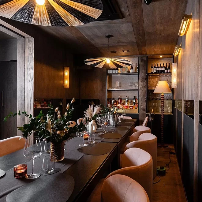 A cosy, elegant dining room with a long table set for a meal. The room features soft, ambient lighting, plush chairs, and decorative plants. Shelves with bottles are visible in the background, adding to the sophisticated atmosphere at SO|LA Soho London, London.