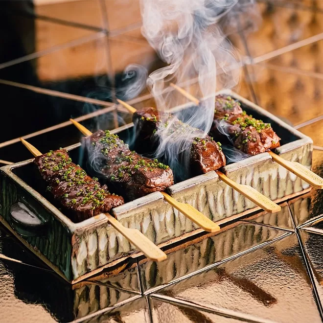 Golden table with a hot plate with 4 pieces of steaming meat on sticks cooking.