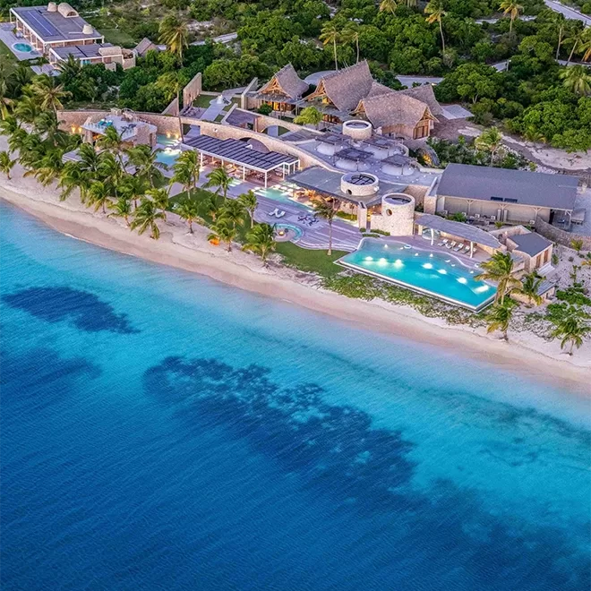 An aerial view of a beach resort with thatched-roof buildings, multiple pools, and palm trees, located directly along a bright turquoise shoreline. at Miavana by Time + Tide, Madagascar.