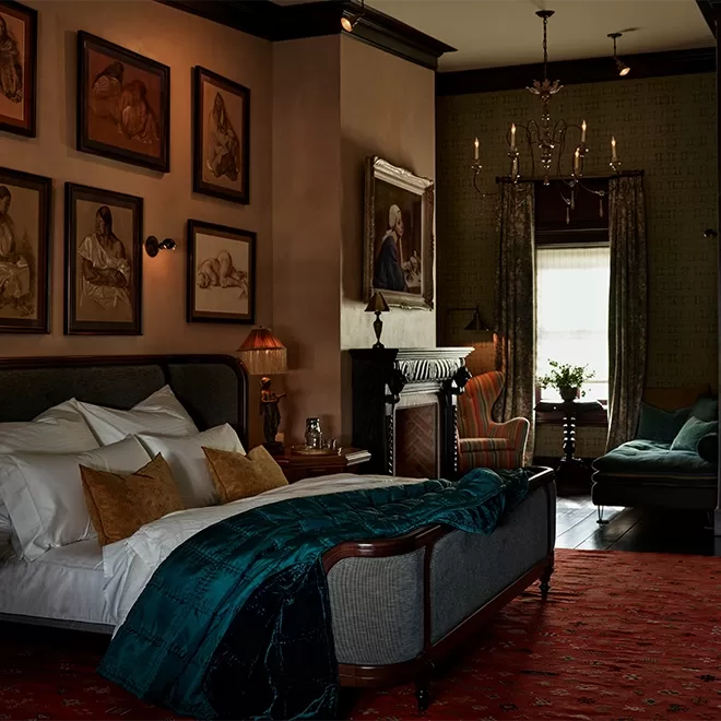 Cosy bedroom with elegant décor featuring a plush bed, teal throw blanket, and red patterned rug. Walls adorned with framed art and a large painting above a fireplace. Armchair and window with curtains provide a warm, inviting ambiance at The Maker Hotel, New York.