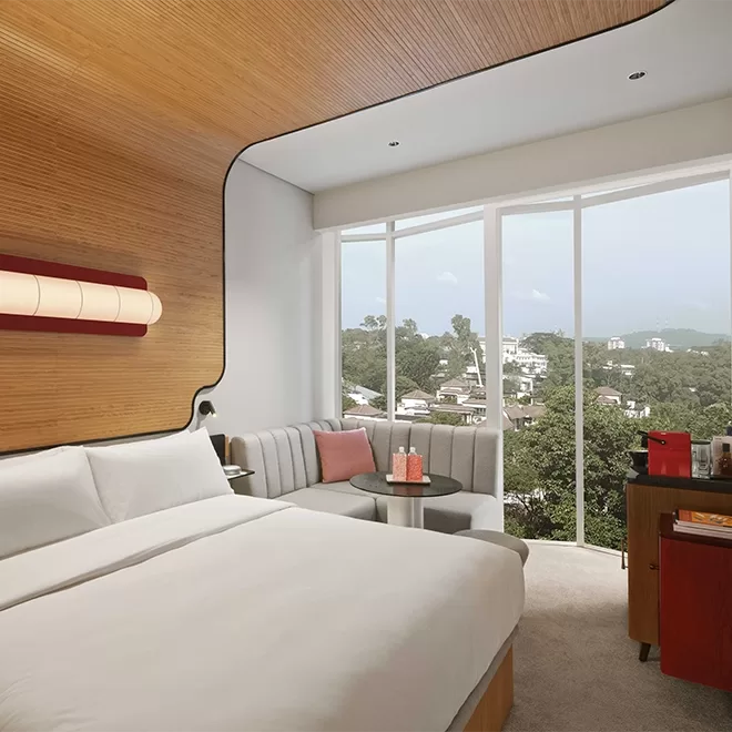Modern hotel room with a large bed, light wood accents, and a cosy seating area by the window. Floor-to-ceiling windows offer a view of a lush green landscape and distant cityscape. A small table and a minibar are present at The Standard, Singapore.