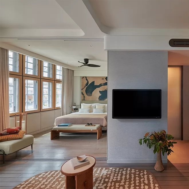 A modern bedroom with a large bed, wooden bench, and abstract wall art. There are large windows with curtains, a ceiling fan, a wall-mounted TV, and a cozy seating area with a patterned rug and small table with flowers at 21 Carpenter hotel in Singapore.