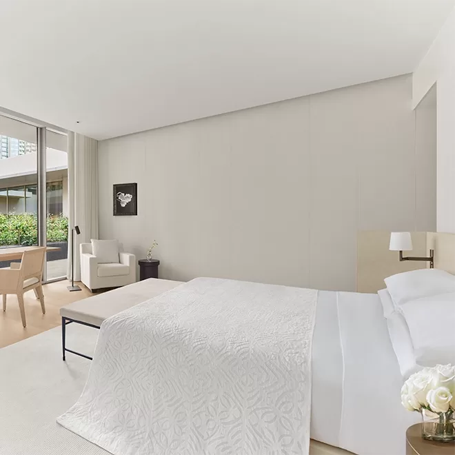 Minimalist bedroom with a large bed covered in a white textured blanket. A chair and small table sit by the window, offering a garden view. The decor includes a black and white wall art piece and a vase with flowers. The room is bright and airy at The Singapore EDITION.