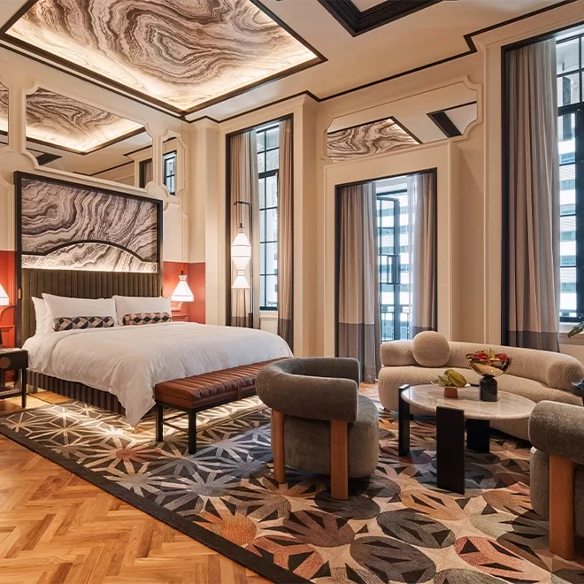 Luxurious hotel room with a large bed, wooden floors, and marble-patterned ceilings. The room features elegant decor, including modern armchairs, a round table, curtains, and a plush rug with geometric designs, offering a cosy and stylish atmosphere at QT Singapore.