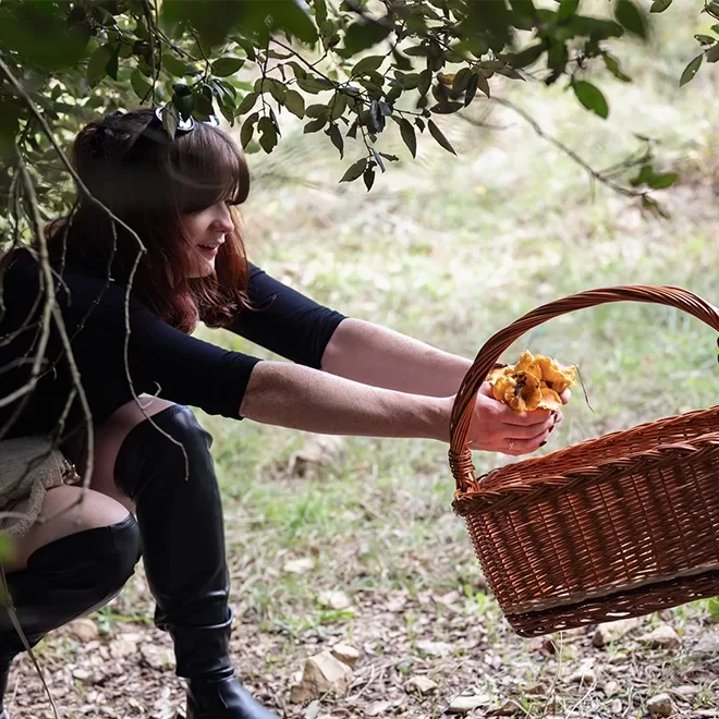 Georgie Young, kneels under trees, collecting mushrooms into a wicker basket. They are dressed in a dark outfit and appear focused on the task. The ground is covered in leaves and grass.