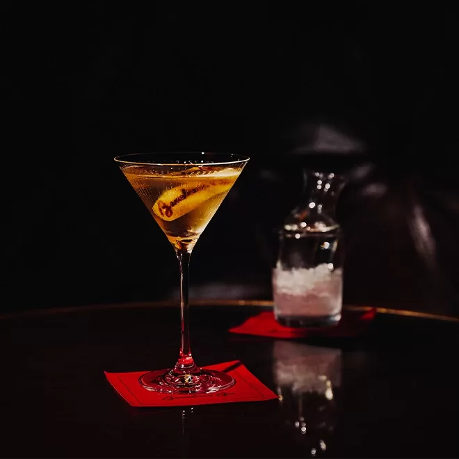 A martini glass with a clear cocktail garnished with lemon twist sits on a red napkin on a dark table. A clear carafe with ice water is in the background, also placed on a red napkin. The setting appears elegant and dimly lit.