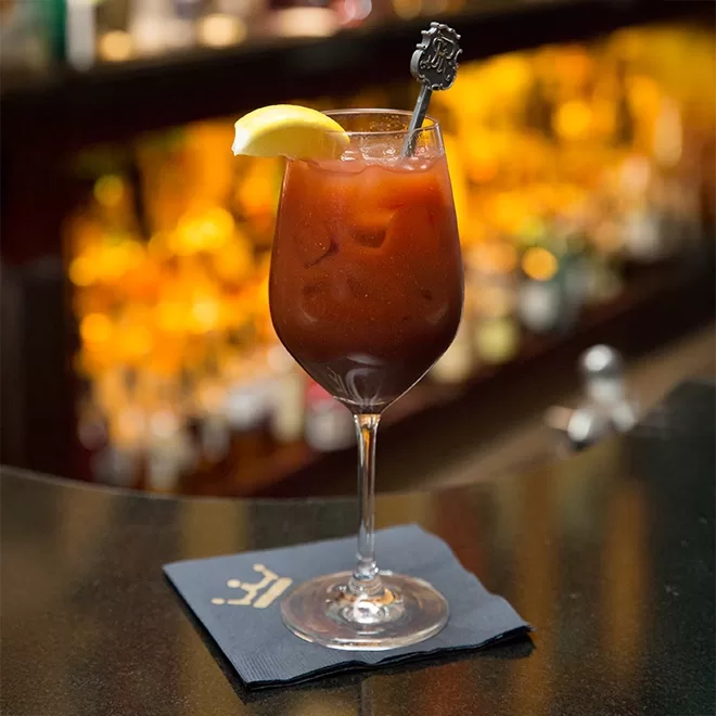 A stemmed glass filled with a dark, red drink garnished with a lemon wedge and a decorative cocktail stirrer. The drink is on a black napkin with a gold emblem. The background is a blurred bar with warm lighting.