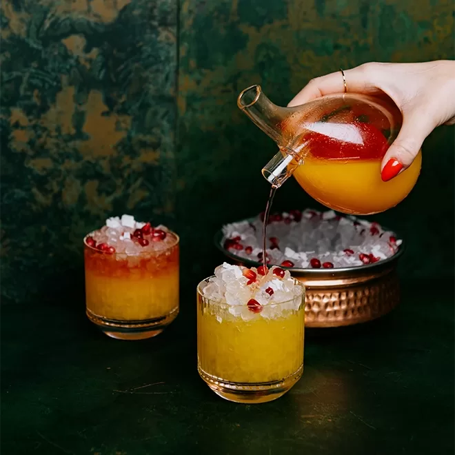 A hand with red nail polish pours a dark liquid from a glass vessel into a cocktail glass filled with ice and orange juice. Another similar cocktail sits nearby. A bowl with ice and pomegranate seeds is in the background on a dark green surface.