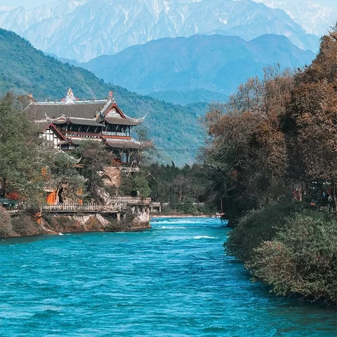 A scenic view of a river winding through lush greenery, with a traditional-style building on the left bank. Majestic mountains rise in the background under a clear blue sky, creating a serene and picturesque landscape.