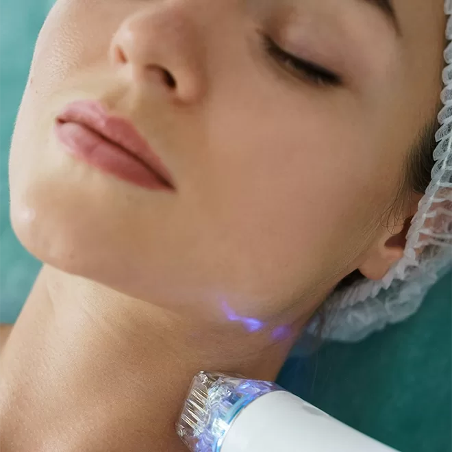 A person receiving a skincare treatment with a device on their neck. The person is lying down with a relaxed expression, wearing a hair cap. The device emits a blue light.