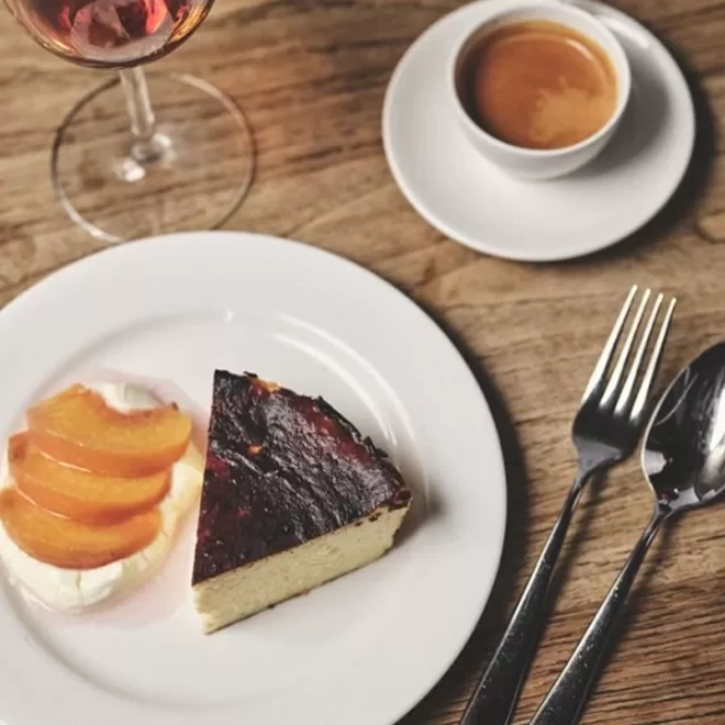 Slice of cheesecake with sliced peaches on whipped cream on a white plate, next to a fork and spoon. A small espresso in a cup and a glass of rosé wine are placed on a wooden table at BRAT Restaurant, Shoreditch.