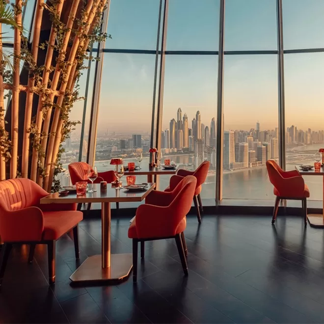 A restaurant in a high up building with beautiful floor to ceiling windows overlooking Dubai and seafront. Tables and chair set up with bamboo along the wall.
