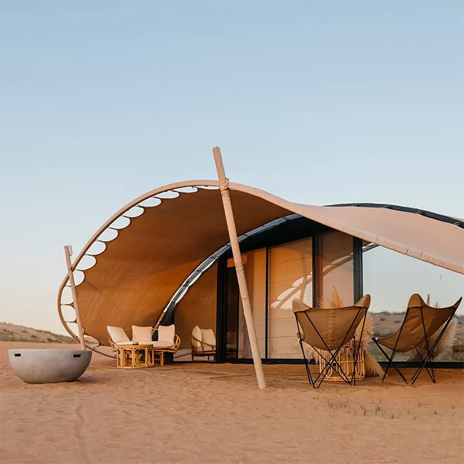 Blue sky and a building on a sandy terrain. The building is dome shaped with double glass doors and a few wicker chairs places outside.