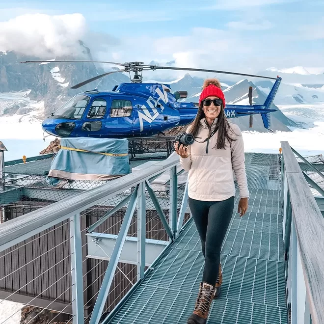 A smiling woman wearing a beanie hat holding a camera, standing on a platform in front of a blue helicopter sat in front of snowy mountains, taking her Sheldon Chalet, Alaska.