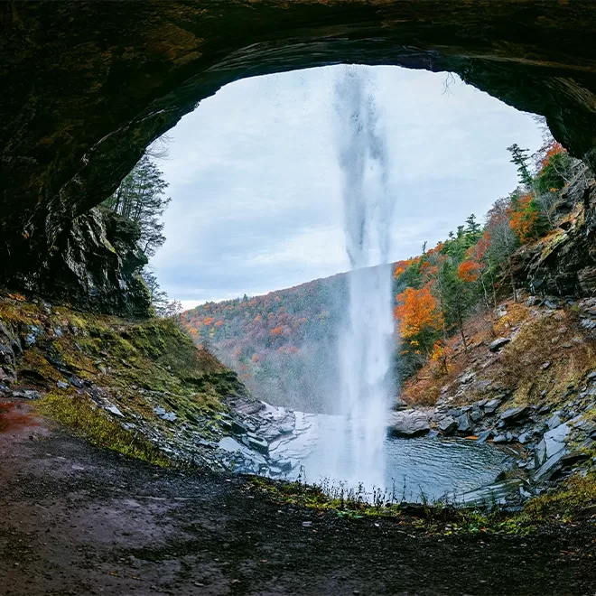 View from behind a waterfall cascading from a cave opening, with a serene pool below. The surrounding landscape is lush with autumn foliage in hues of orange and red, under an overcast sky.