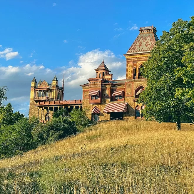 A historic mansion with ornate brickwork and multiple towers reminiscent of the Olana State Historic Site stands on a grassy hill under a blue sky with scattered clouds. Trees surround the hillside, partially shading the building.