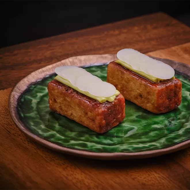 Two rectangular pieces of winter radish cake, vegetable treacle, and tarragon are artistically arranged on a textured green plate set on a wooden surface. Served at new restaurant AngloThai in London.
