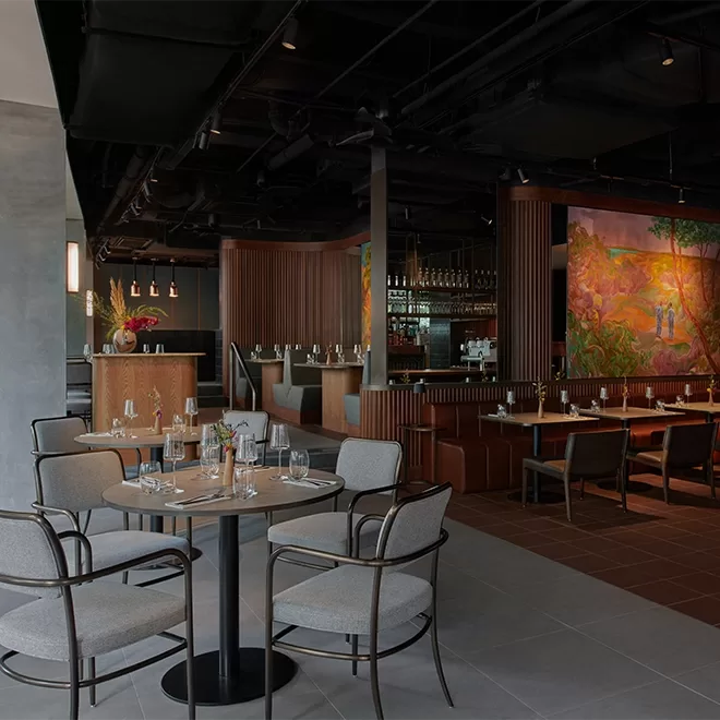 A modern restaurant interior with a cosy atmosphere, featuring several tables set with glassware and napkins. The space includes elegant upholstered chairs, a mural on the wall, and a bar area in the background with ambient lighting at 21 Carpenter hotel in Singapore.