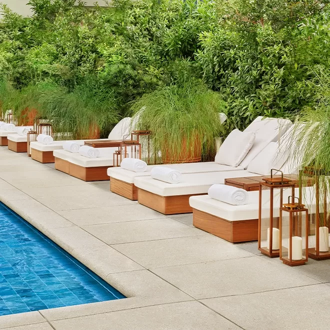 A peaceful poolside setting with a row of white cushioned lounge chairs, each with a rolled towel. Lush green plants provide a backdrop, while lanterns sit beside the chairs on wooden side tables, creating a serene atmosphere at The Singapore EDITION.