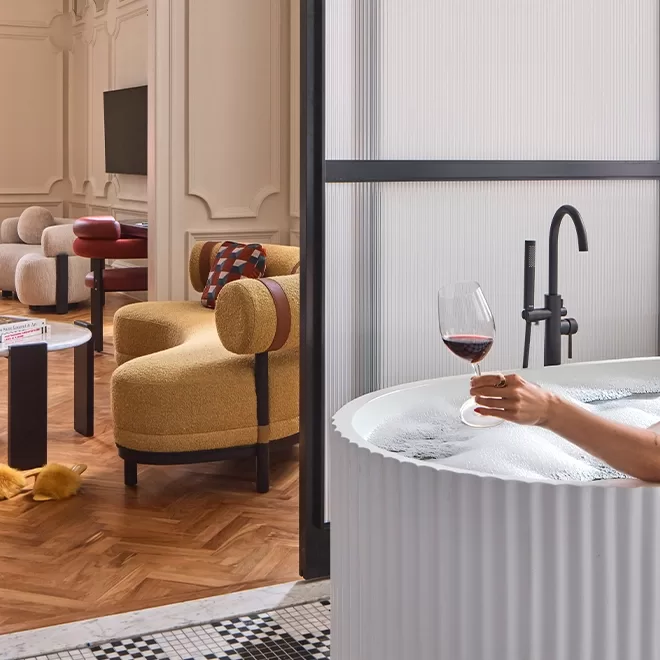 A luxurious bathroom scene features a person relaxing in a modern white bathtub with a glass of red wine. The room includes elegant decor with plush, curved yellow seating and a stylish wooden floor.