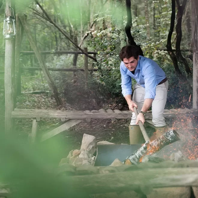 A man in a blue shirt uses a large wooden stick to tend a fire pit outdoors, surrounded by green foliage and trees. He appears focused on managing the flames as he bends over the fire.